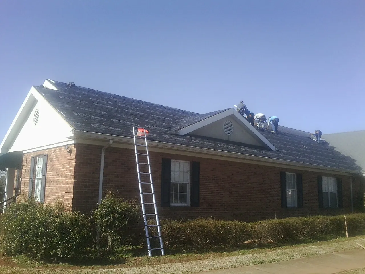 Crew tearing shingles off a brick ranch roof with a ladder set — Metal Roof Repair work by our crew in Quaker Hill