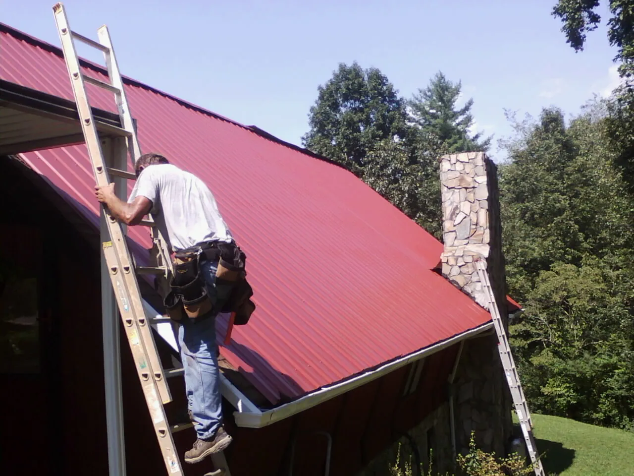 Roofer on a ladder beside a red metal roof and stone chimney — Soffit & Fascia Repair by Roofing Brothers in Quaker Hill